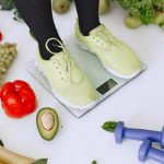 Person standing on scales surrounded by fresh produce and fitness equipment symbolizing a balanced lifestyle.