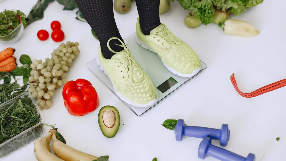 Person standing on scales surrounded by fresh produce and fitness equipment symbolizing a balanced lifestyle.